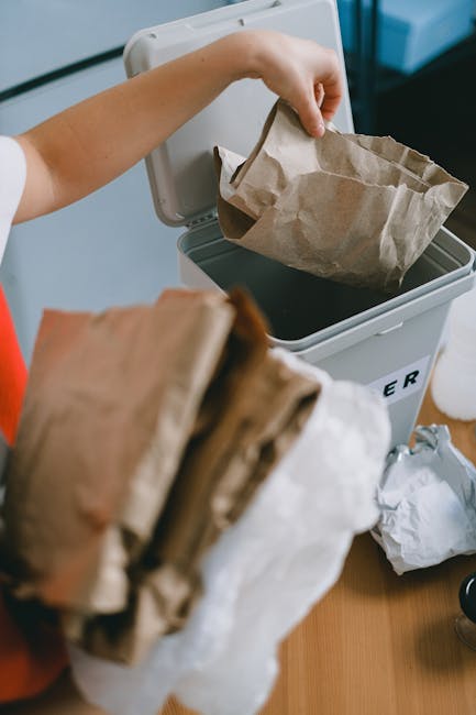 Close-up image of a black plastic rubbish bag filled with household waste, positioned in the foreground against a plain, light-colored wall. The bag is tightly secured at the top with visible folds an