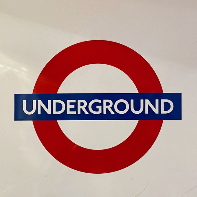 A long, curved underground London Underground station platform featuring white tiled walls with the red and blue London Underground roundel sign prominently displayed on the left. The platform has a d
